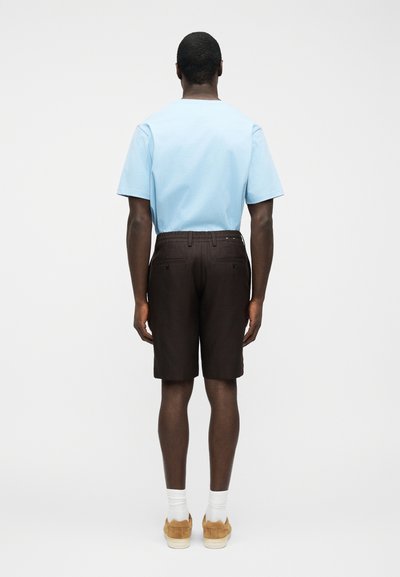 Homme de dos portant une chemise à manches courtes bleu clair, un short marron foncé jusqu'aux genoux, des chaussettes blanches et des baskets en daim beige sur fond blanc.