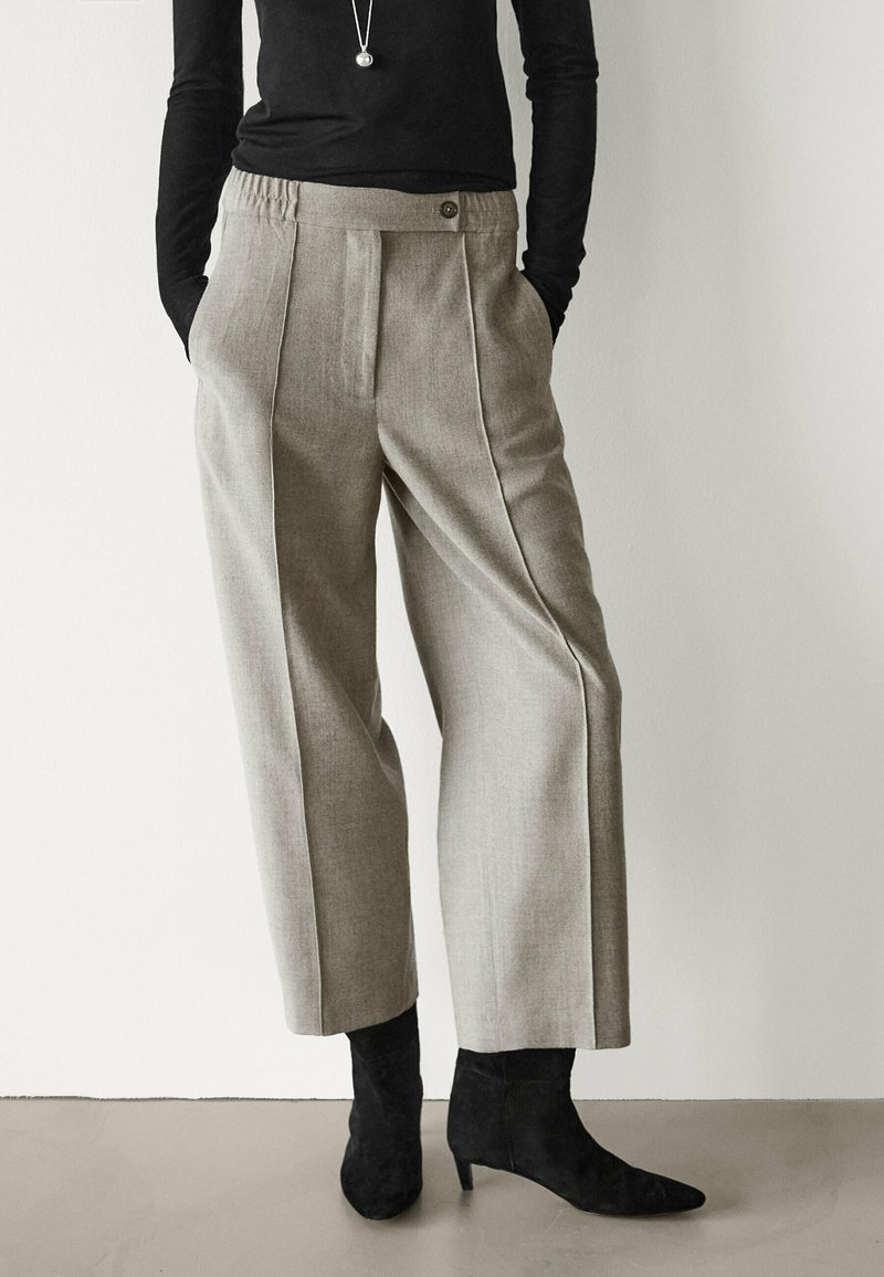 Pantalon gris ajusté à coupe droite, avec plis et bouton à la taille. Associé à un haut noir ajusté et des bottines noires à bout pointu.