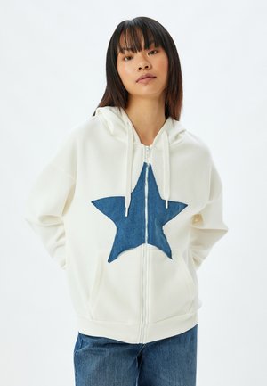 White zip-up hoodie featuring a blue denim star on the front, with a hood, drawstrings, and a relaxed fit. Worn over blue jeans.