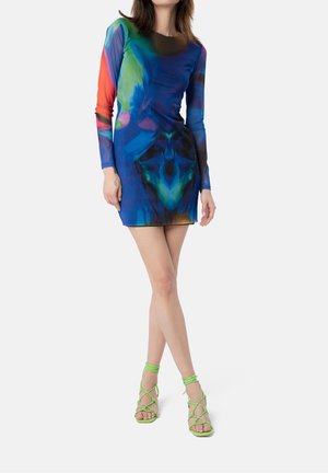 Robe mini à manches longues colorée avec des motifs abstraits bleus, verts et oranges ; associée à des sandales à talons aiguilles vertes.