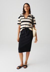 Gestreiftes Strickoberteil in Creme und Schwarz, mit V-Ausschnitt und kurzen Ärmeln; kombiniert mit einem schwarzen Midirock mit elastischem Bund und schwarzen Sandalen.