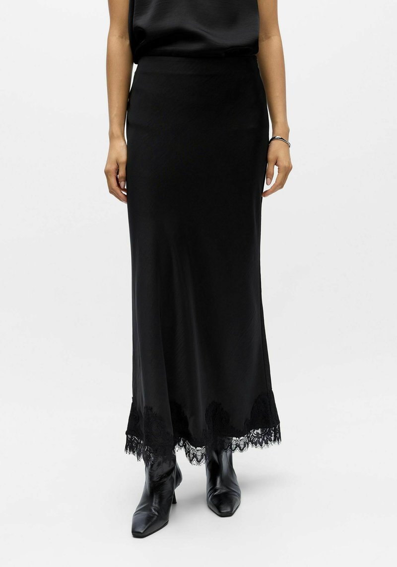 Jupe maxi en satin noir avec une ceinture ajustée et un détail en dentelle délicate à l'ourlet, accompagnée de bottines noires fines à la cheville.