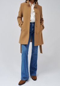 Cappotto in misto lana cammello con collo alto e chiusura frontale con bottoni, abbinato a jeans a zampa di colore azzurro chiaro e stivaletti in camoscio marrone.