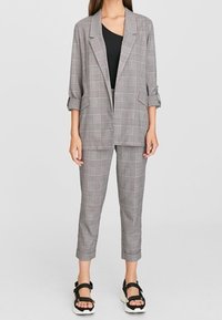 Femme portant un blazer à carreaux gris et un pantalon court assorti, avec un haut noir et des sandales noires à bout ouvert avec semelles blanches.