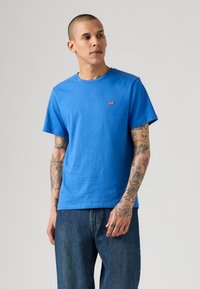 Άνδρας με ολοκληρωμένα τατουάζ στα χέρια, φορώντας μπλε μπλουζάκι Levi's και μπλε τζιν, κοιτάζει στο πλάι σε έναν απλό φόντο.