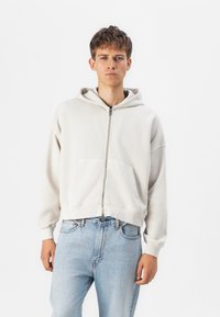 Haut à capuche zippé gris clair avec une poche frontale, coupe décontractée et cordon de serrage à la capuche. Associé à un jean en denim bleu clair. Texture douce.