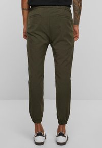 Olijfgroene tapered pants met een elastische tailleband, twee achterzakken en elastische boorden, gecombineerd met witte sneakers met zwarte accenten.