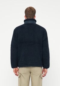 Muškarac u tamnoplavoj flis jakni s visokim ovratnikom i bež cargo hlačama, prikazan s leđa na običnoj bijeloj pozadini.