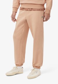 Pantaloni da tuta color pesca realizzati in tessuto morbido, con una cintura a motivo, polsini elasticizzati e un piccolo dettaglio con logo verde sul lato.