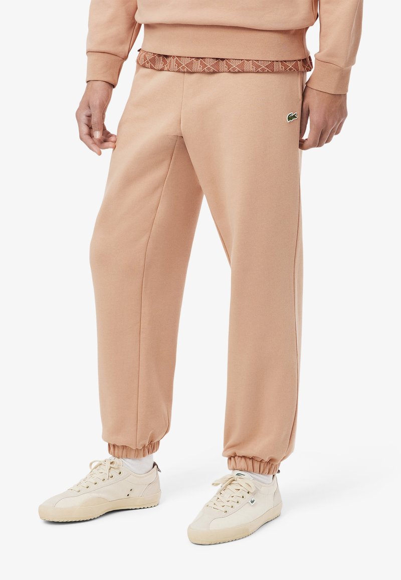 Pantaloni da tuta color pesca realizzati in tessuto morbido, con una cintura a motivo, polsini elasticizzati e un piccolo dettaglio con logo verde sul lato.