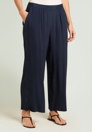 Vrouw draagt loszittende marineblauwe wijde broek met zijzakken, gecombineerd met donkerbruine teenslippers op een lichte achtergrond.
