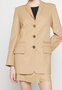 Blazer beige ajusté avec un revers cranté, trois boutons et deux poches avant ; présente des accents de couture horizontale près des poches.