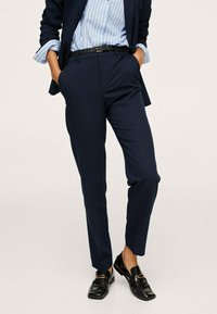 Osoba ubrana w granatowe, dopasowane spodnie, czarny pasek, czarne mokasyny oraz niebieską koszulę w paski z podwiniętymi rękawami.