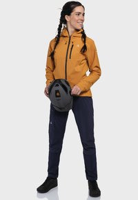 Frau in senfgelber Jacke, dunklen Hosen und schwarzen Schuhen, die einen schwarzen Fahrradhelm hält, zur Seite schaut und lächelt.