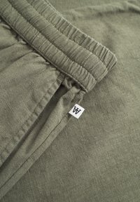 Olivgrüner Leinenstoff mit einem gerafften Taillenbund und einem kleinen weißen Etikett mit "W." Texturierte Oberfläche mit einem weichen Finish.