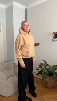 Sweatshirt bege claro com bolso frontal e cordão, combinado com calças pretas de perna larga e sapatos pretos robustos. Ambiente interior com plantas.