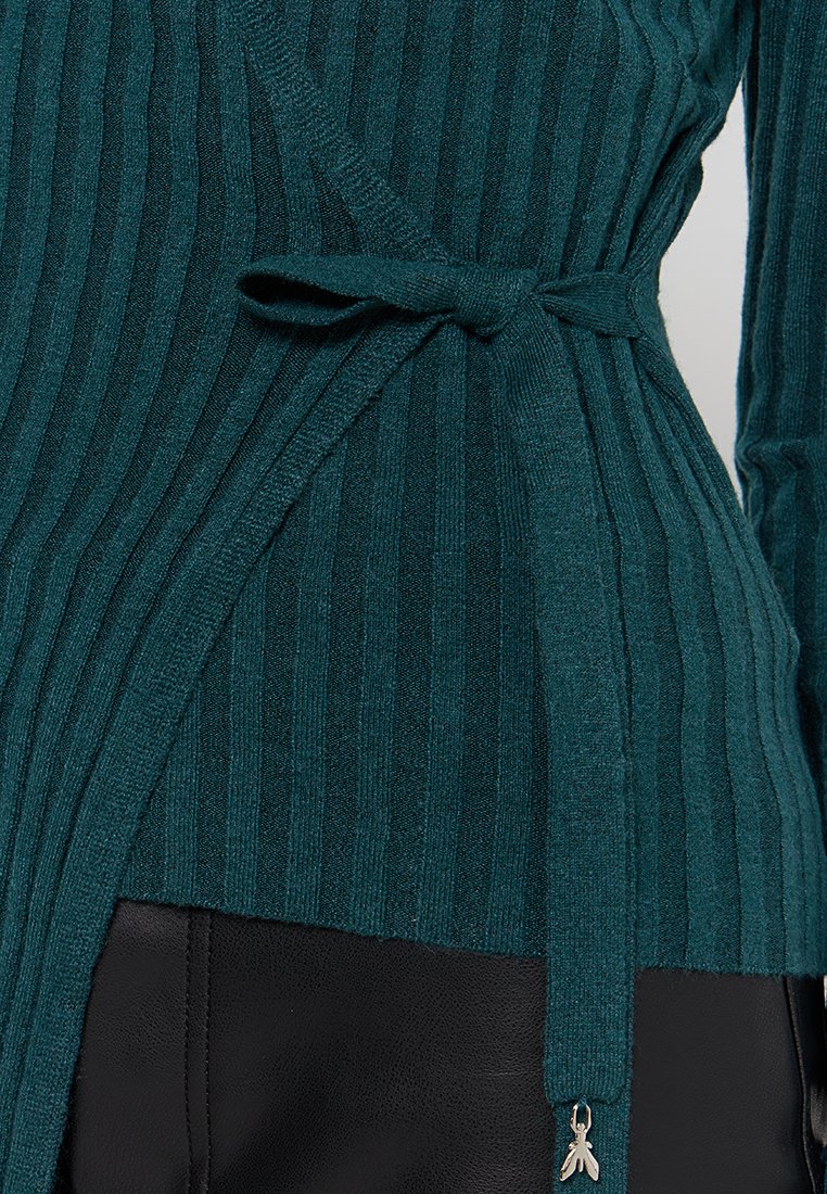 Maglione avvolgente a coste color turchese legato a fiocco in vita sopra pantaloni in pelle nera con un piccolo charm a forma di insetto in argento all'estremità del nodo.
