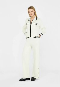 Conjunto deportivo de punto blanco con detalles en negro. Incluye una chaqueta con cremallera y pantalones acampanados con botones en el dobladillo. Textura suave.