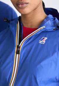 Chaqueta ligera azul con capucha, con cierre de cremallera y acentos de rayas multicolores. Logo de K-WAY en el lado izquierdo del pecho.