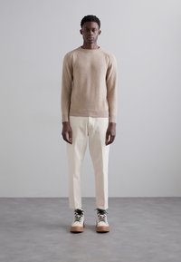 Suéter de punto beige con dobladillo acanalado, pantalones blancos a medida y botas crema con cordones negros. El modelo está de pie frente a un fondo gris liso.