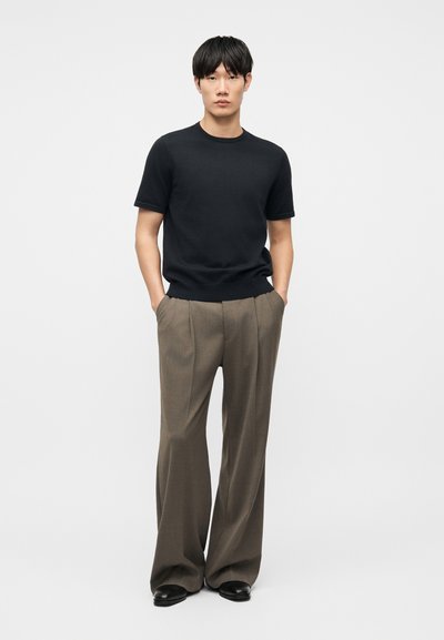 Νεαρός άνδρας στέκεται με τα χέρια στις τσέπες, φορώντας μαύρο πουκάμισο με κοντά μανίκια, χαλαρό καφέ παντελόνι και μαύρα παπούτσια σε απλό φόντο.