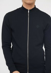 Sweatshirt zippé noir pour homme avec un col haut, des poignets côtelés, des poches avant et un logo circulaire sur la poitrine. Tissu doux avec une texture lisse.
