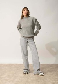 Maglione grigio chiaro lavorato a maglia con collo alto, caratterizzato da una texture a coste e motivi sottili, abbinato a jeans grigio chiaro a zampa e sneaker.