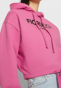 Sudadera corta rosa fabricada con tela suave, con capucha ajustable y texto negro "FIORUCCI MILANO" en el frente.