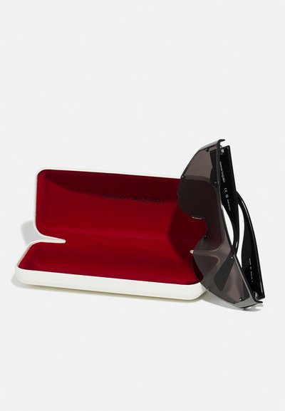 Occhiali da sole neri con un design elegante e angolare, dotati di lenti in gradienti di nero e grigio, custoditi in un astuccio rigido bianco e rosso.