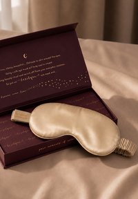 Seidengoldfarbene Augenmaske, die auf einer offenen dunkelvioletten Schachtel mit goldenem Schriftzug auf beigem Stoffhintergrund liegt.