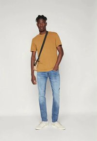 T-shirt de algodão bege, ajuste slim, combinada com jeans de denim azul claro. O acessório inclui uma pequena bolsa com uma alça preta. Os sapatos são ténis brancos.