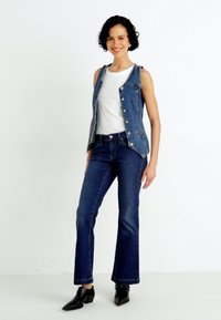 Gilet in denim con bottoni argentati sopra una camicia bianca, abbinato a jeans a zampa d'elefante blu scuro e stivaletti neri alla caviglia. Design semplice e pulito.