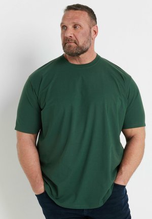 Muskelbepackter Mann mit Bart trägt ein dunkelgrünes kurzärmeliges Hemd, steht mit den Händen in den Taschen und blickt nach links vor einem einfarbigen Hintergrund.