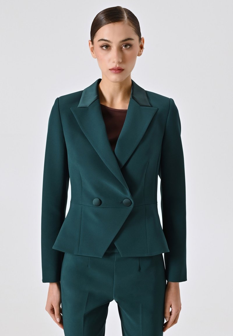 Blazer doppiopetto teal in tessuto strutturato con vita a peplo, colletto stondato e pantaloni coordinati, caratterizzati da linee pulite e bottoni.