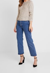 Kobieta w beżowym swetrze w cętki, niebieskich jeansach z wysokim stanem i czarnych szpilkach z paskiem wokół kostki stoi na tle jednolitego tła.