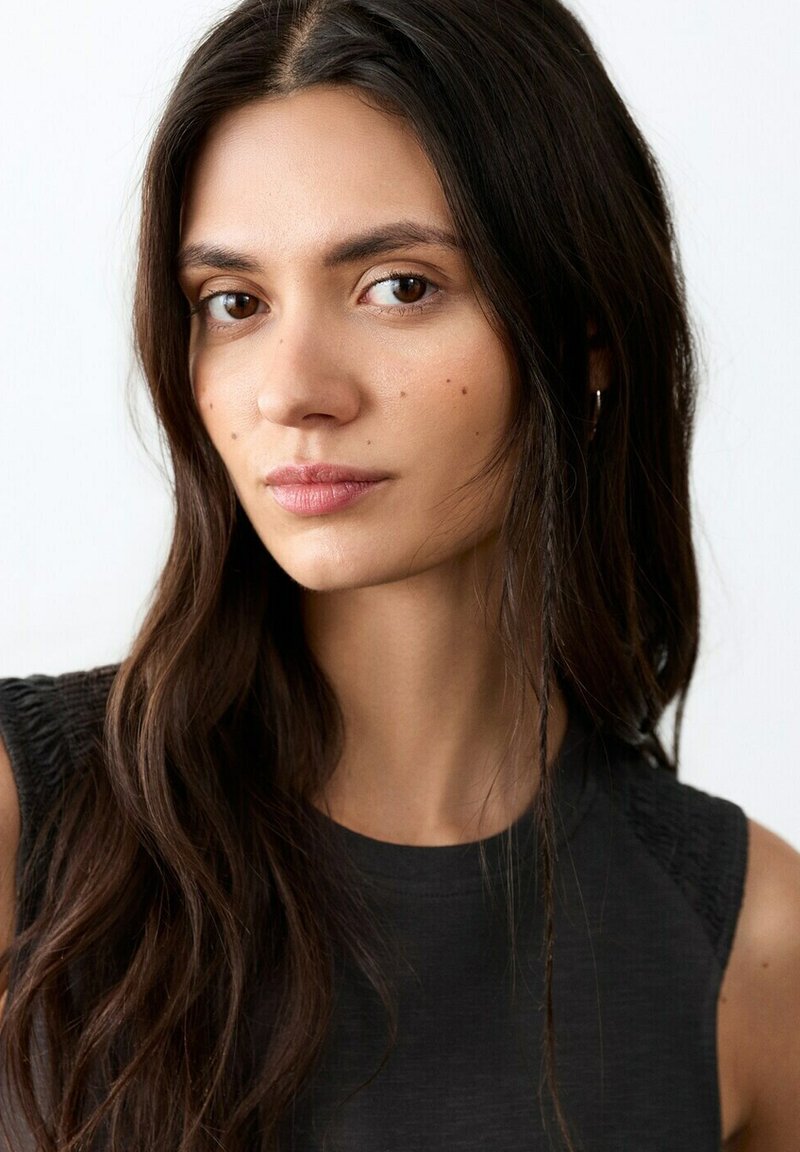 Mlajša ženska z dolgimi temnimi lasmi, oblečena v črno majico brez rokavov, premišljeno gleda v kamero na enostavni beli podlagi.