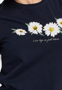 Chemise bleu marine avec des marguerites blanches et des feuilles vertes sur la poitrine avec le texte "Live life in full bloom" en italique.