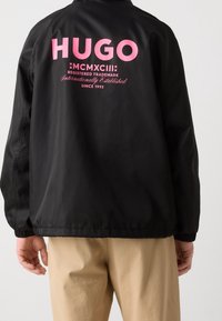 Czarna kurtka z wysokim kołnierzem, z dużym różowym napisem "HUGO" i dodatkowymi oznaczeniami z tyłu. Elastyczne mankiety i dół.