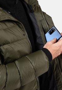 Oliwkowa kurtka puchowa z pikowanym wzorem, otwarta z przodu, z wewnętrzną kieszenią trzymającą niebieskiego smartfona. Ubranie posiada czarny dzianinowy kołnierz.