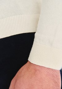 Jasnokremowy sweter w dzianinę z prążkowanymi mankietami, zbliżenie na rękaw, na ciemnym tle.
