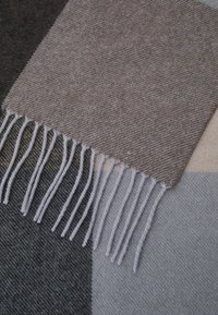 Wollendecke mit geometrischem Muster in Braun, Grau und Beige. Sie hat eine strukturierte Oberfläche und geknotete Fransenränder.