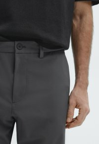 Pantalons gris sur mesure avec une texture lisse, dotés d'une fermeture à bouton et à zip classique, ainsi que de passants de ceinture. Le design inclut des poches latérales.