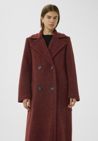 Cappotto doppiopetto in misto lana bordeaux profondo, con grandi bottoni neri, ampi revers e tasche laterali; maniche lunghe e testurizzate.
