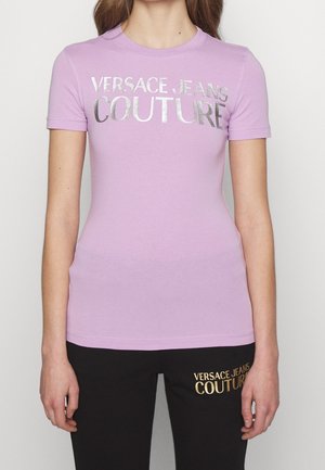 Camiseta de manga corta lila de tejido suave con impresión metálica plateada "VERSACE JEANS COUTURE" en el frente. Combinada con pantalones de chándal negros con logo dorado.