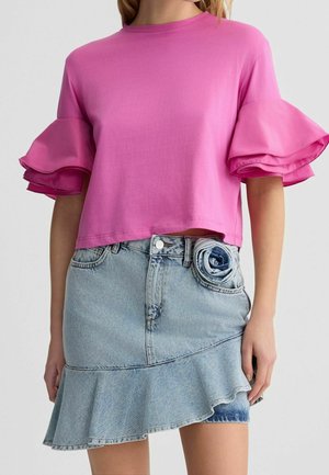 Donna che indossa una maglia a maniche corte di un rosa acceso con maniche arricciate e una gonna di jeans asimmetrica azzurro chiaro con un dettaglio a fiore di jeans arrotolato.