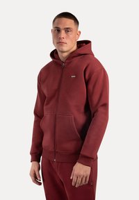 Felpa rossa con zip realizzata in tessuto morbido, dotata di tasca a marsupio, polsini a coste e cappuccio. Piccolo logo sul petto.