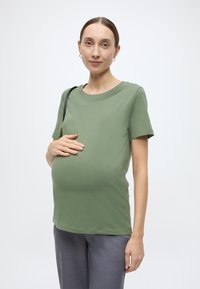 Κοντομάνικη μπλούζα εγκυμοσύνης σε απαλό ελιάς πράσινο ύφασμα, με κοντά μανίκια και στρογγυλή λαιμόκοψη, φορεμένη με γκρι παντελόνι. Το μοντέλο αγγίζει απαλά την κοιλιά της.