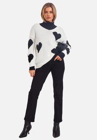 Weißer Strickpullover mit schwarzen Herzmustern, kombiniert mit schwarzen, engen Hosen. Das Model trägt schwarze Stiefeletten mit Absatz.