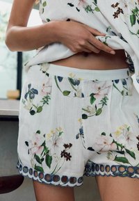 Weiße Blumenshorts aus leichtem Material, mit blauem gewelltem Saum und aufwendigen Blumenmustern in Pink, Gelb und Grün.