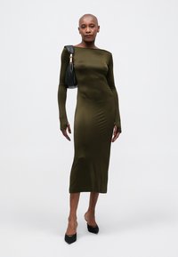 Olive grøn color ilga rankovė su prigludusiu siluetu, turinti valtinį iškirpimą. Modelis laiko juodą rankinę, apsirengęs juodais smailiais bateliais.